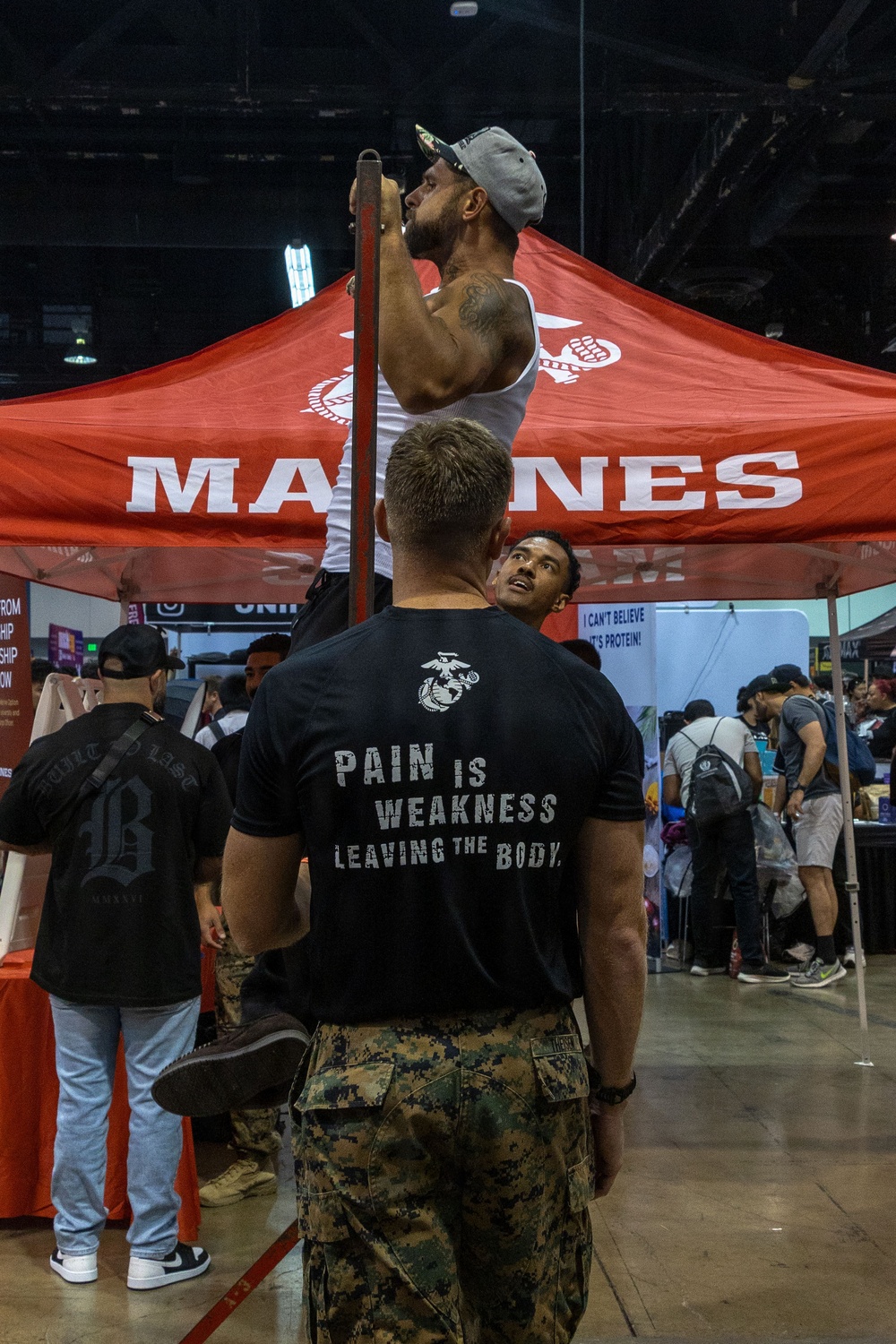 The image shows a U.S. Marines recruiting booth at what appears to be an indoor expo or fitness convention. A man in a white tank top is performing pull-ups on a bar attached to the red Marines-branded tent, while a Marine in uniform observes. The setting includes multiple vendor booths and a crowd of attendees in the background.