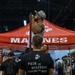 The image shows a U.S. Marines recruiting booth at what appears to be an indoor expo or fitness convention. A man in a white tank top is performing pull-ups on a bar attached to the red Marines-branded tent, while a Marine in uniform observes. The setting includes multiple vendor booths and a crowd of attendees in the background.