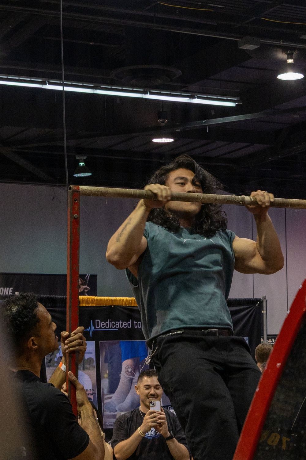 A muscular man with long dark hair is performing a pull-up or chin-up on a horizontal bar mounted on a red metal frame structure, likely at a fitness expo or public demonstration event. He is wearing a teal sleeveless shirt and black pants, with his chin at bar level. Spectators below are watching and recording the performance on a smartphone.