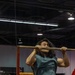 A muscular man with long dark hair is performing a pull-up or chin-up on a horizontal bar mounted on a red metal frame structure, likely at a fitness expo or public demonstration event. He is wearing a teal sleeveless shirt and black pants, with his chin at bar level. Spectators below are watching and recording the performance on a smartphone.