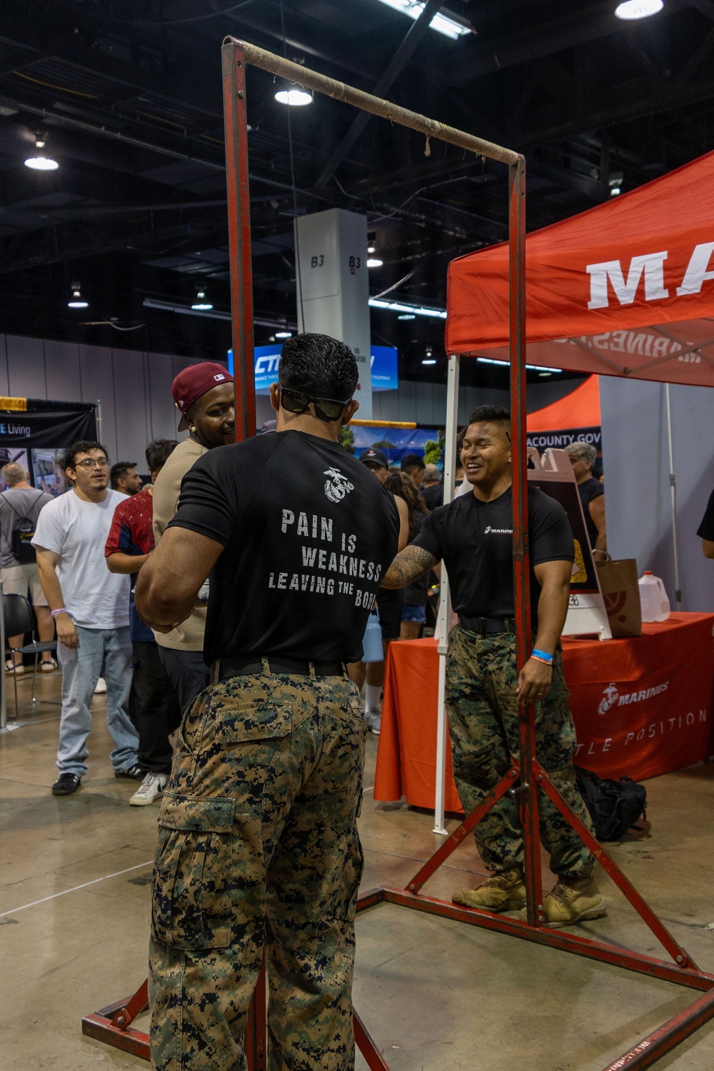 A U.S. Marines recruiting booth at what appears to be a career or community expo held in a large convention center. Two individuals in Marine Corps MARPAT camouflage uniforms are interacting near a pull-up bar station, with one wearing a black t-shirt reading 'Pain is Weakness Leaving the Body' with the Eagle, Globe, and Anchor emblem. A red Marines-branded canopy tent and promotional table are visible in the background.