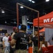 A U.S. Marines recruiting booth at what appears to be a career or community expo held in a large convention center. Two individuals in Marine Corps MARPAT camouflage uniforms are interacting near a pull-up bar station, with one wearing a black t-shirt reading 'Pain is Weakness Leaving the Body' with the Eagle, Globe, and Anchor emblem. A red Marines-branded canopy tent and promotional table are visible in the background.