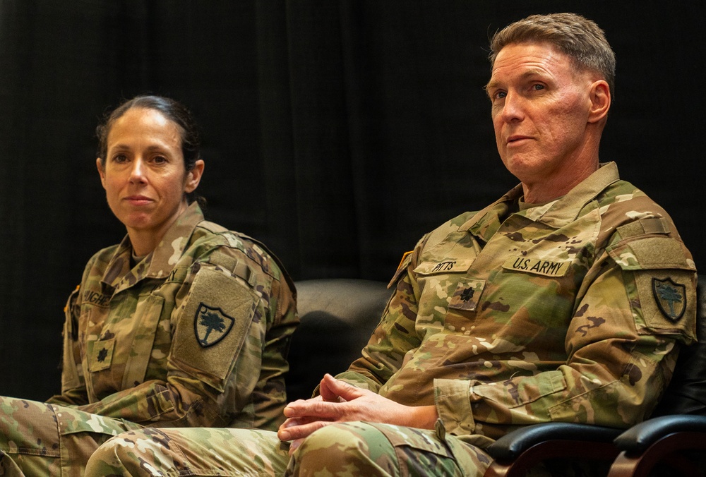 Two U.S. Army soldiers in Operational Camouflage Pattern (OCP) uniforms are seated side by side against a dark backdrop, appearing to be attending or participating in a formal military event or briefing. The soldier on the right has a visible name tape reading 'PITTS' and 'U.S. ARMY' tape, along with a gold oak leaf cluster rank insignia indicating the rank of Lieutenant Colonel. Both soldiers wear the same unit patch on their left shoulders, identifiable as the 218th Maneuver Enhancement Brigade (South Carolina National Guard palmetto tree patch).
