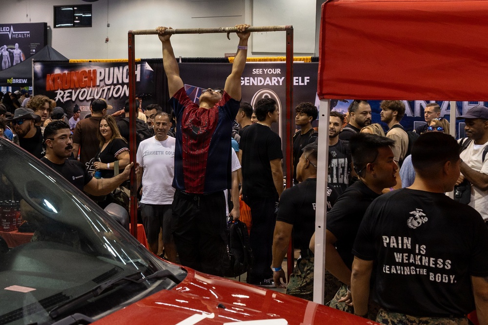 A fitness or health expo event where a person is performing pull-ups on a freestanding pull-up bar surrounded by a large crowd of spectators. Several vendor booths are visible in the background, including 'Krunch Punch Revolution' and a motivational banner. A red vehicle and a red canopy tent are visible in the foreground, along with individuals wearing U.S. Marine Corps shirts.
