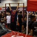A fitness or health expo event where a person is performing pull-ups on a freestanding pull-up bar surrounded by a large crowd of spectators. Several vendor booths are visible in the background, including 'Krunch Punch Revolution' and a motivational banner. A red vehicle and a red canopy tent are visible in the foreground, along with individuals wearing U.S. Marine Corps shirts.
