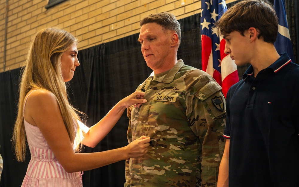 A U.S. Army soldier in OCP (Operational Camouflage Pattern) uniform is having an insignia or pin affixed to his chest by a young woman in a pink striped dress, while a teenage boy looks on. This appears to be a military promotion or award ceremony taking place indoors, likely in a gymnasium or assembly hall. American flags and what appears to be a unit flag are visible in the background.