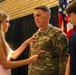 A U.S. Army soldier in OCP (Operational Camouflage Pattern) uniform is having an insignia or pin affixed to his chest by a young woman in a pink striped dress, while a teenage boy looks on. This appears to be a military promotion or award ceremony taking place indoors, likely in a gymnasium or assembly hall. American flags and what appears to be a unit flag are visible in the background.