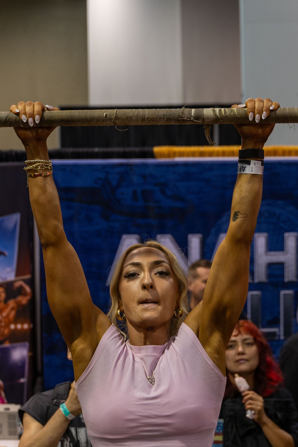 A muscular blonde woman is performing a dead hang or pull-up on a worn wooden/wrapped bar at what appears to be a fitness expo or competition. She is wearing a light pink sleeveless top, gold jewelry, and wristbands, with visible muscle definition in her arms and shoulders. A blue banner partially reading 'ANA' and 'H' is visible in the background along with spectators.