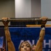 A muscular blonde woman is performing a dead hang or pull-up on a worn wooden/wrapped bar at what appears to be a fitness expo or competition. She is wearing a light pink sleeveless top, gold jewelry, and wristbands, with visible muscle definition in her arms and shoulders. A blue banner partially reading 'ANA' and 'H' is visible in the background along with spectators.