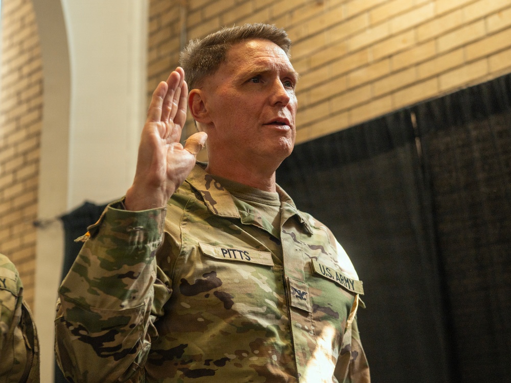 A U.S. Army soldier with the surname 'Pitts' is raising his right hand, appearing to take an oath or be sworn in during a military ceremony. He is wearing the Army Combat Uniform (ACU) in MultiCam/OCP pattern with a Colonel's eagle insignia visible on his chest. The setting appears to be an indoor military facility with brick walls and a dark curtain backdrop.