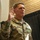 Lt. Col. Hughes and Lt. Col. Pitts promotion ceremony to colonel A U.S. Army soldier with the surname 'Pitts' is raising his right hand, appearing to take an oath or be sworn in during a military ceremony. He is wearing the Army Combat Uniform (ACU) in MultiCam/OCP pattern with a Colonel's eagle insignia visible on his chest. The setting appears to be an indoor military facility with brick walls and a dark curtain backdrop.