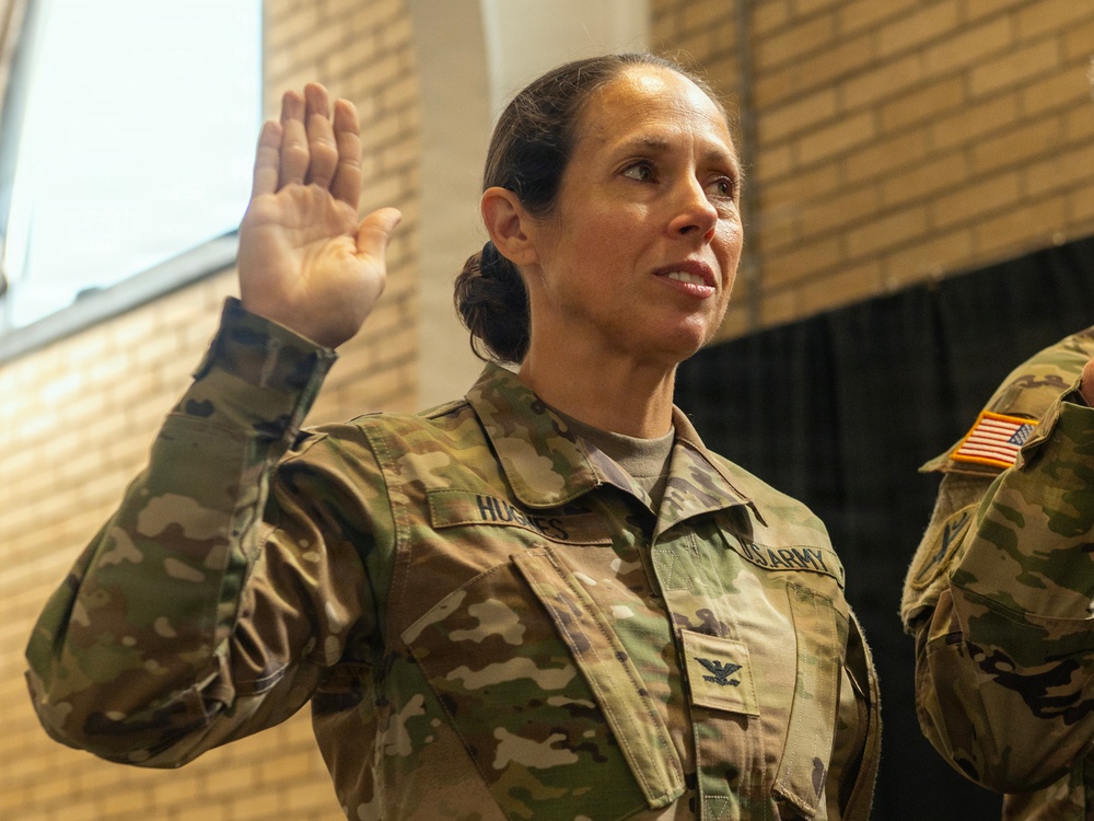 A U.S. Army Colonel is raising her right hand, appearing to take an oath or be sworn in during a military ceremony. Her uniform displays the name tape 'HUGHES' (partially visible) and an eagle insignia indicating the rank of Colonel (O-6). A second uniformed soldier is partially visible to the right.