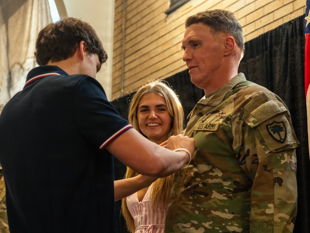 A U.S. Army soldier in OCP (Operational Camouflage Pattern) uniform is having an insignia or pin affixed to his chest by a young man, while a smiling young woman assists or observes closely. This appears to be a military promotion or award ceremony. The setting is an indoor venue with brick walls and dark draping in the background.