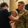 Lt. Col. Hughes and Lt. Col. Pitts promotion ceremony to colonel A U.S. Army soldier in OCP (Operational Camouflage Pattern) uniform is having an insignia or pin affixed to his chest by a young man, while a smiling young woman assists or observes closely. This appears to be a military promotion or award ceremony. The setting is an indoor venue with brick walls and dark draping in the background.