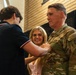 A U.S. Army soldier in OCP (Operational Camouflage Pattern) uniform is having an insignia or pin affixed to his chest by a young man, while a smiling young woman assists or observes closely. This appears to be a military promotion or award ceremony. The setting is an indoor venue with brick walls and dark draping in the background.