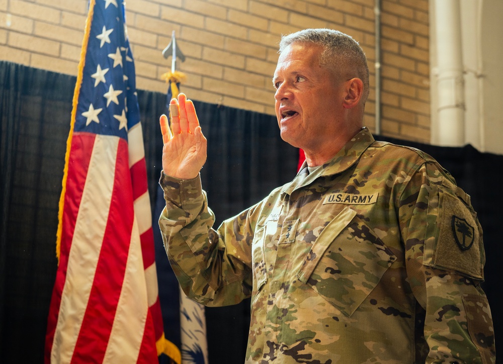 A U.S. Army soldier in OCP (Operational Camouflage Pattern) uniform is raising his right hand, appearing to take an oath or pledge. An American flag and what appears to be a state flag are visible in the background. The setting appears to be an indoor military ceremony in a brick-walled facility.