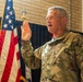 A U.S. Army soldier in OCP (Operational Camouflage Pattern) uniform is raising his right hand, appearing to take an oath or pledge. An American flag and what appears to be a state flag are visible in the background. The setting appears to be an indoor military ceremony in a brick-walled facility.