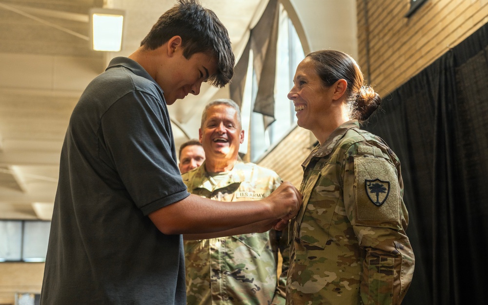 A young civilian male is pinning an insignia or rank device onto the uniform of a female U.S. Army soldier during what appears to be a military promotion or award ceremony. Two other uniformed U.S. Army soldiers are visible in the background, smiling and observing the moment. The setting appears to be an indoor facility such as a gymnasium or event hall.