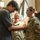 Lt. Col. Hughes and Lt. Col. Pitts promotion ceremony to colonel A young civilian male is pinning an insignia or rank device onto the uniform of a female U.S. Army soldier during what appears to be a military promotion or award ceremony. Two other uniformed U.S. Army soldiers are visible in the background, smiling and observing the moment. The setting appears to be an indoor facility such as a gymnasium or event hall.
