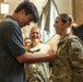 A young civilian male is pinning an insignia or rank device onto the uniform of a female U.S. Army soldier during what appears to be a military promotion or award ceremony. Two other uniformed U.S. Army soldiers are visible in the background, smiling and observing the moment. The setting appears to be an indoor facility such as a gymnasium or event hall.