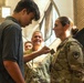 A young civilian male is pinning a rank insignia or badge onto a female U.S. Army soldier in military camouflage (OCP) uniform during what appears to be a promotion or award ceremony. A smiling male soldier in the background observes the event. The setting appears to be an indoor military or civic facility.