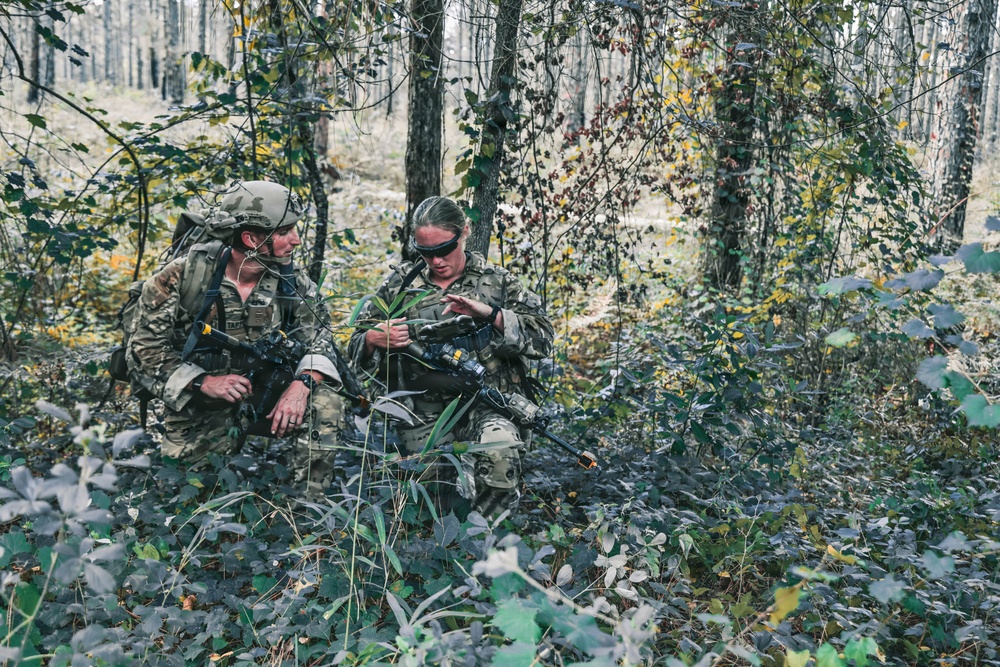 Two soldiers in full camouflage gear are positioned in a forested area, appearing to review information or equipment together. Both are armed and equipped with tactical gear, headsets, and backpacks, suggesting an active field operation or military training exercise. The woodland setting with mixed foliage provides natural concealment.