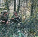 Two soldiers in full camouflage gear are positioned in a forested area, appearing to review information or equipment together. Both are armed and equipped with tactical gear, headsets, and backpacks, suggesting an active field operation or military training exercise. The woodland setting with mixed foliage provides natural concealment.