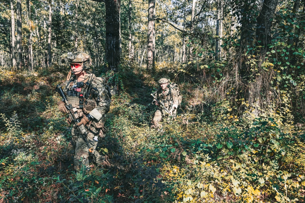 Two soldiers in full multicam camouflage gear are conducting a patrol through a dense woodland area. The foreground soldier displays a visible US flag patch on their left shoulder and is equipped with a rifle, plate carrier, and red-tinted protective eyewear. The image appears to depict a military training exercise or operational patrol in a temperate forest environment.