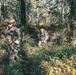 Two soldiers in full multicam camouflage gear are conducting a patrol through a dense woodland area. The foreground soldier displays a visible US flag patch on their left shoulder and is equipped with a rifle, plate carrier, and red-tinted protective eyewear. The image appears to depict a military training exercise or operational patrol in a temperate forest environment.