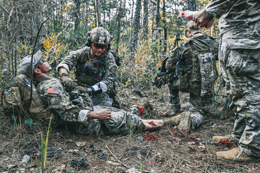 The image depicts what appears to be a military combat casualty care training exercise in a wooded environment. Several soldiers in U.S. Army MultiCam/OCP camouflage uniforms are attending to a simulated casualty on the ground. The scene includes simulated wound makeup on the casualty's leg, consistent with a field medical training scenario.