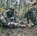 The image depicts what appears to be a military combat casualty care training exercise in a wooded environment. Several soldiers in U.S. Army MultiCam/OCP camouflage uniforms are attending to a simulated casualty on the ground. The scene includes simulated wound makeup on the casualty's leg, consistent with a field medical training scenario.