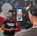 A camera operator wearing headphones is filming a woman standing in front of a mural that reads 'WELDER'. The subject, wearing a dark t-shirt and yellow safety glasses, is visible both in the background (slightly out of focus) and sharply on the camera's external monitor/field display. This appears to be a professional video production shoot, likely documenting a welding or vocational training program.