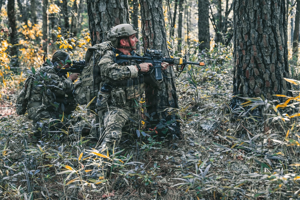 Two individuals in full military-style camouflage gear are positioned behind large tree trunks in a forested environment, holding rifles in a tactical stance. The foreground figure is crouching and aiming forward while the background figure appears to be in a lower prone-adjacent position also aiming a weapon. The setting suggests a military training exercise or airsoft/tactical simulation scenario, indicated by the orange muzzle tip visible on the foreground rifle.
