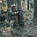 Two individuals in full military-style camouflage gear are positioned behind large tree trunks in a forested environment, holding rifles in a tactical stance. The foreground figure is crouching and aiming forward while the background figure appears to be in a lower prone-adjacent position also aiming a weapon. The setting suggests a military training exercise or airsoft/tactical simulation scenario, indicated by the orange muzzle tip visible on the foreground rifle.
