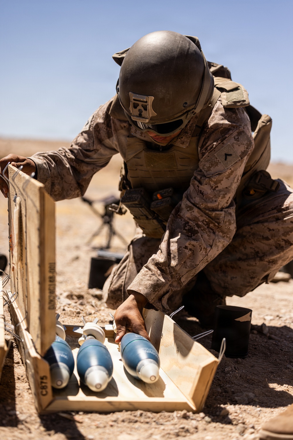 A military service member in desert camouflage (MARPAT pattern) and combat gear is handling blue-painted mortar rounds from a wooden ammunition crate in an arid, desert environment. The rounds appear to be inert training munitions based on their blue color coding, which is standard NATO designation for practice/training ordnance. A mortar tube is visible in the background, suggesting an active training exercise.