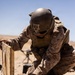 A military service member in desert camouflage (MARPAT pattern) and combat gear is handling blue-painted mortar rounds from a wooden ammunition crate in an arid, desert environment. The rounds appear to be inert training munitions based on their blue color coding, which is standard NATO designation for practice/training ordnance. A mortar tube is visible in the background, suggesting an active training exercise.
