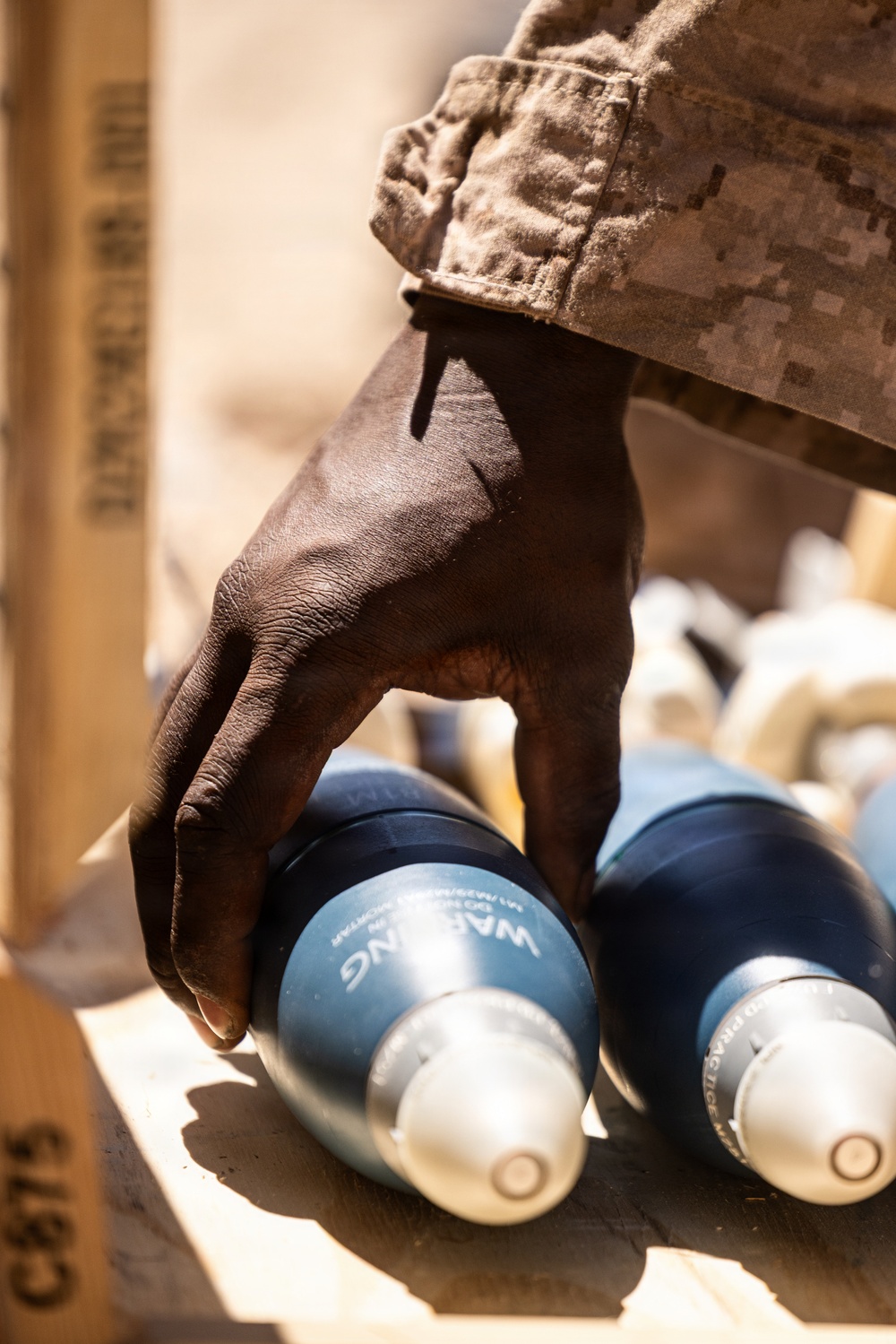 A person wearing desert digital camouflage uniform is handling blue-painted practice mortar rounds stored in a wooden crate. The rounds are visibly marked with 'WARNING' and 'PRACTICE' text, indicating these are inert training munitions. The close-up photograph captures the handling of what appear to be M929/M930-type 60mm or 81mm practice mortar rounds.