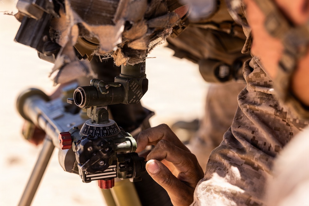A soldier in desert camouflage uniform is adjusting the sight/aiming mechanism on what appears to be a mortar system. The close-up shot focuses on the soldier's hands manipulating a numbered elevation dial and optical sight assembly. The setting appears to be an arid, desert environment based on the sandy background.