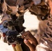 A soldier in desert camouflage uniform is adjusting the sight/aiming mechanism on what appears to be a mortar system. The close-up shot focuses on the soldier's hands manipulating a numbered elevation dial and optical sight assembly. The setting appears to be an arid, desert environment based on the sandy background.