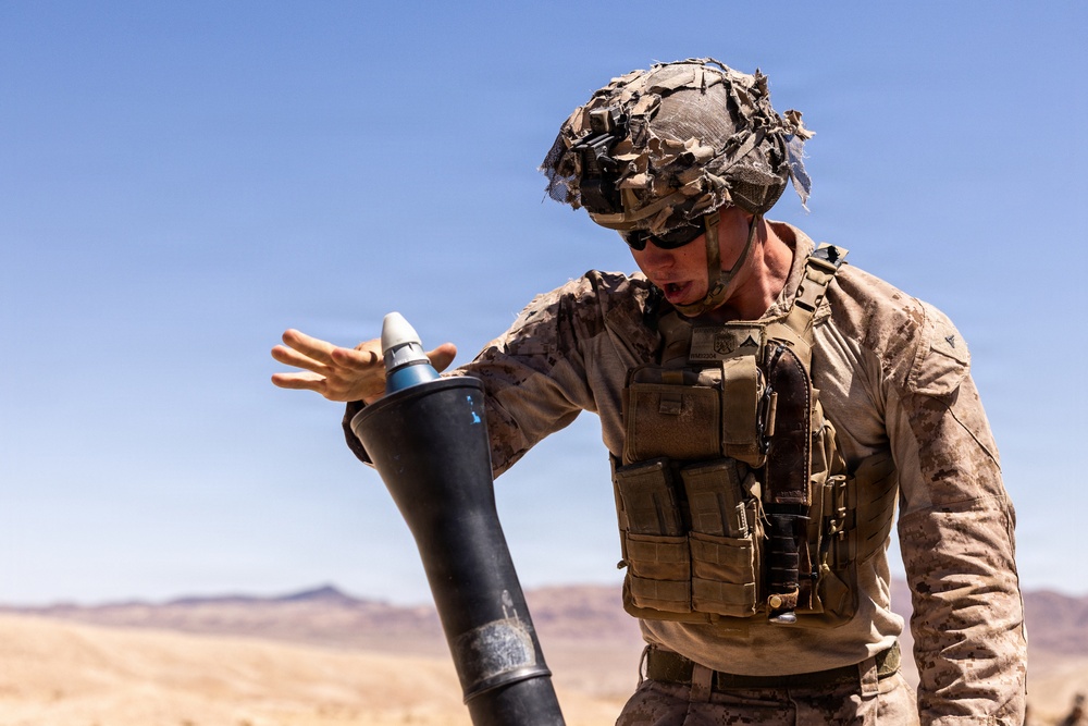A military service member in desert camouflage uniform and tactical gear is handling a mortar round over a mortar tube in an arid, desert environment. The soldier appears to be in the process of loading or preparing to drop the round into the tube. The setting and uniform suggest a training exercise or combat operation in a desert region.