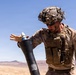 A military service member in desert camouflage uniform and tactical gear is handling a mortar round over a mortar tube in an arid, desert environment. The soldier appears to be in the process of loading or preparing to drop the round into the tube. The setting and uniform suggest a training exercise or combat operation in a desert region.