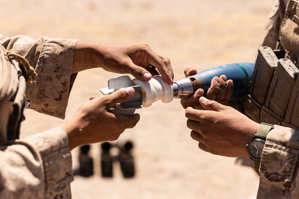 Two individuals in desert camouflage military uniforms are handling what appears to be a mortar round or training munition outdoors in an arid environment. The image is a close-up shot focused on their hands assembling or inspecting the munition components. Additional munitions or equipment are visible in the blurred background.