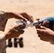 Two individuals in desert camouflage military uniforms are handling what appears to be a mortar round or training munition outdoors in an arid environment. The image is a close-up shot focused on their hands assembling or inspecting the munition components. Additional munitions or equipment are visible in the blurred background.