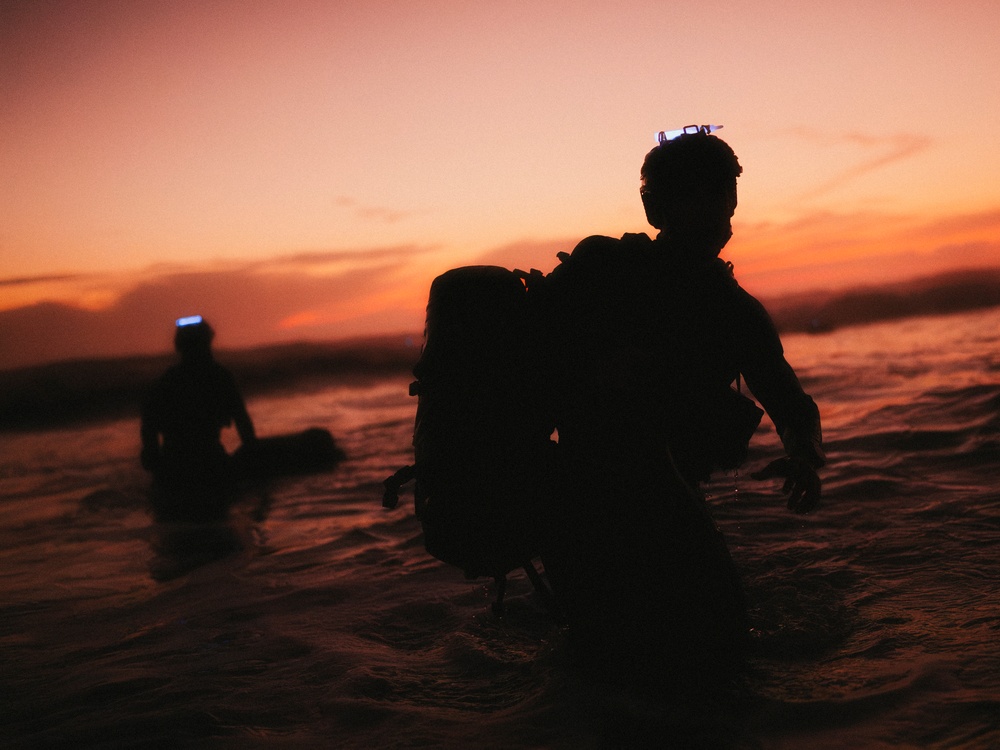 Two silhouetted figures are wading through shallow ocean water during a vivid orange and red sunset. Both individuals are wearing large backpacks and appear to have headlamps or helmet-mounted equipment. The dramatic backlighting creates strong silhouettes against the colorful sky.