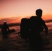Two silhouetted figures are wading through shallow ocean water during a vivid orange and red sunset. Both individuals are wearing large backpacks and appear to have headlamps or helmet-mounted equipment. The dramatic backlighting creates strong silhouettes against the colorful sky.