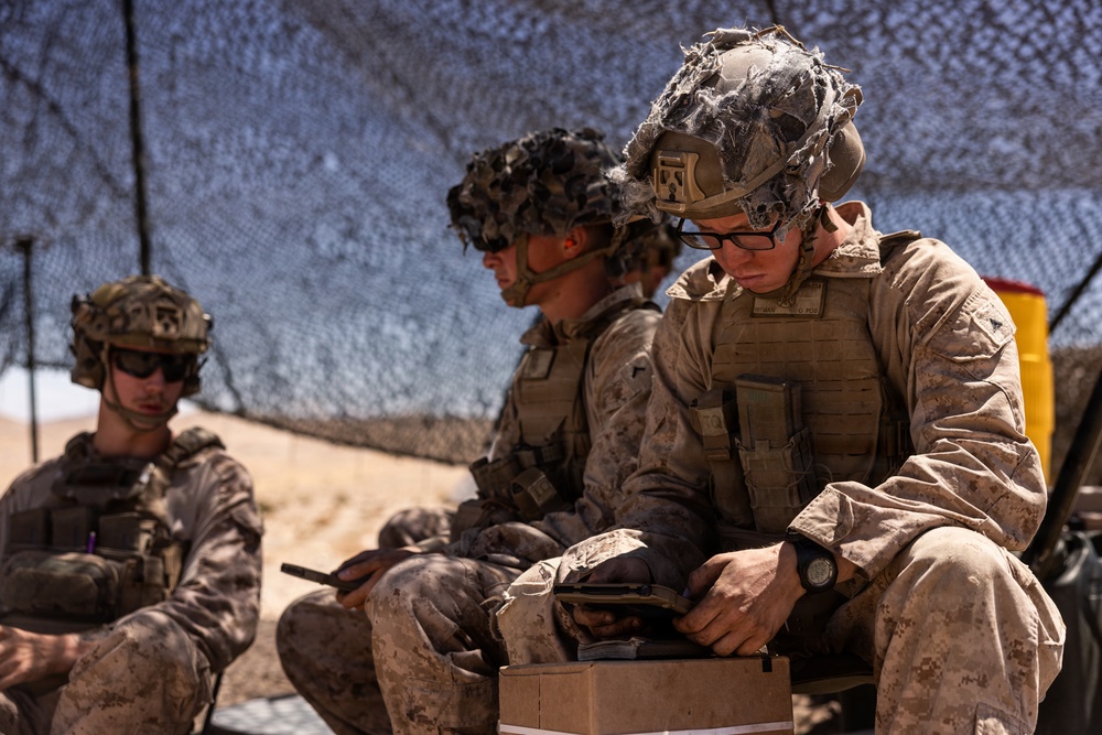 Three military personnel in desert camouflage uniforms are seated under a camouflage netting in an arid environment. The foreground soldier, wearing glasses and a tactical helmet with netting, appears to be inspecting or handling equipment on a small box. The scene suggests a field operation or training exercise in a desert setting.