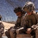 Three military personnel in desert camouflage uniforms are seated under a camouflage netting in an arid environment. The foreground soldier, wearing glasses and a tactical helmet with netting, appears to be inspecting or handling equipment on a small box. The scene suggests a field operation or training exercise in a desert setting.