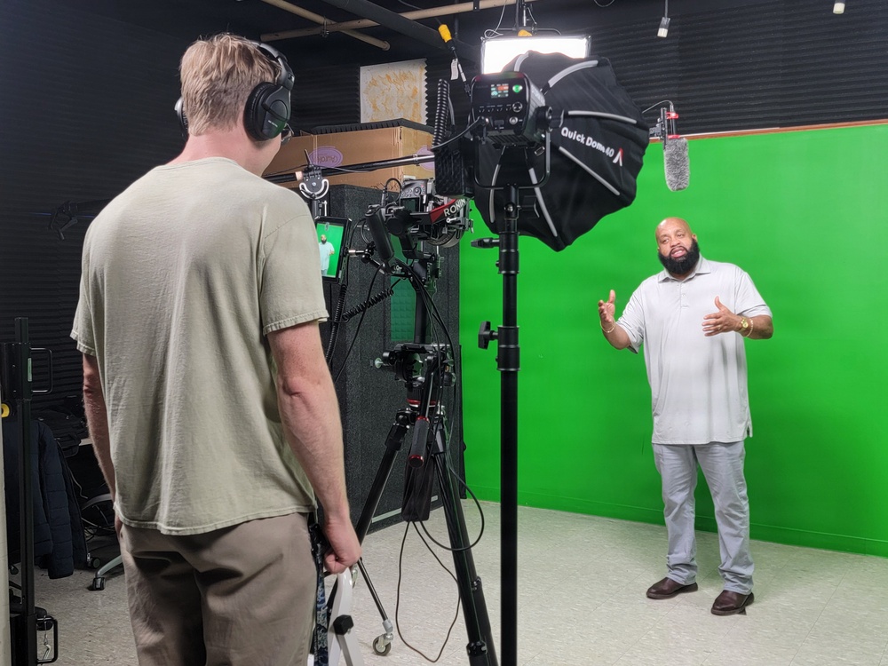 A professional video production shoot is taking place inside a studio with a green screen backdrop. One person stands in front of the green screen as a subject/talent, gesturing with their hands, while a camera operator manages equipment in the foreground. The setup includes professional lighting, a gimbal-mounted camera, and a boom microphone.