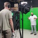 A professional video production shoot is taking place inside a studio with a green screen backdrop. One person stands in front of the green screen as a subject/talent, gesturing with their hands, while a camera operator manages equipment in the foreground. The setup includes professional lighting, a gimbal-mounted camera, and a boom microphone.
