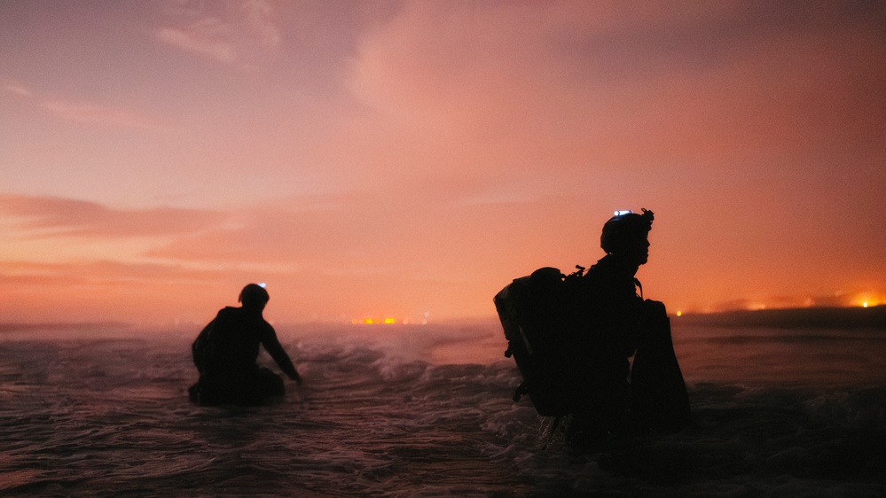 Two silhouetted figures are wading through ocean surf during twilight or nighttime conditions against a vivid orange-red sky. Both individuals are wearing tactical gear including helmets, backpacks, and what appear to be headlamps or night-vision mounts. Distant city lights are visible on the horizon, suggesting a coastal urban environment nearby.
